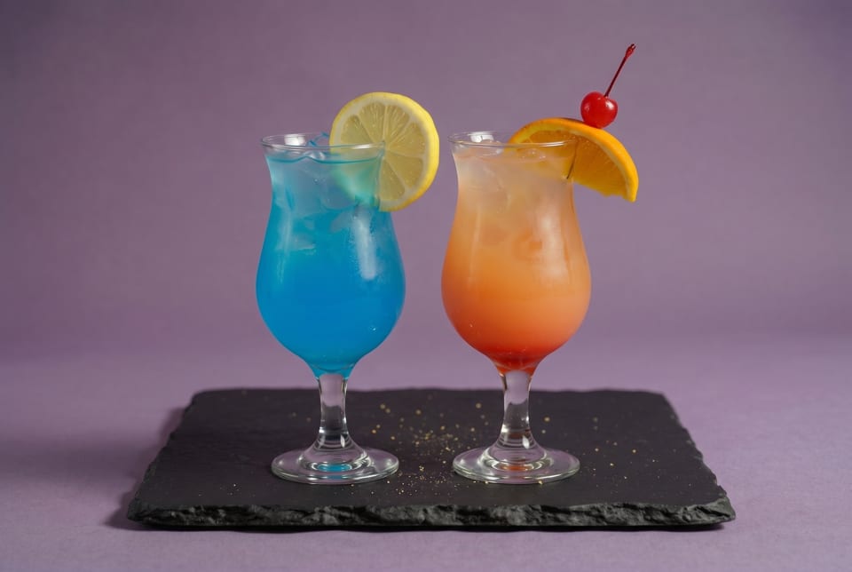 Two colorful cocktails, one blue with lemon and one orange with cherry and orange slice, on a slate board.