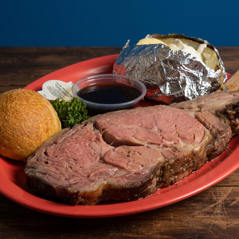 A prime rib dinner with a baked potato, roll, and au jus on a red plate.