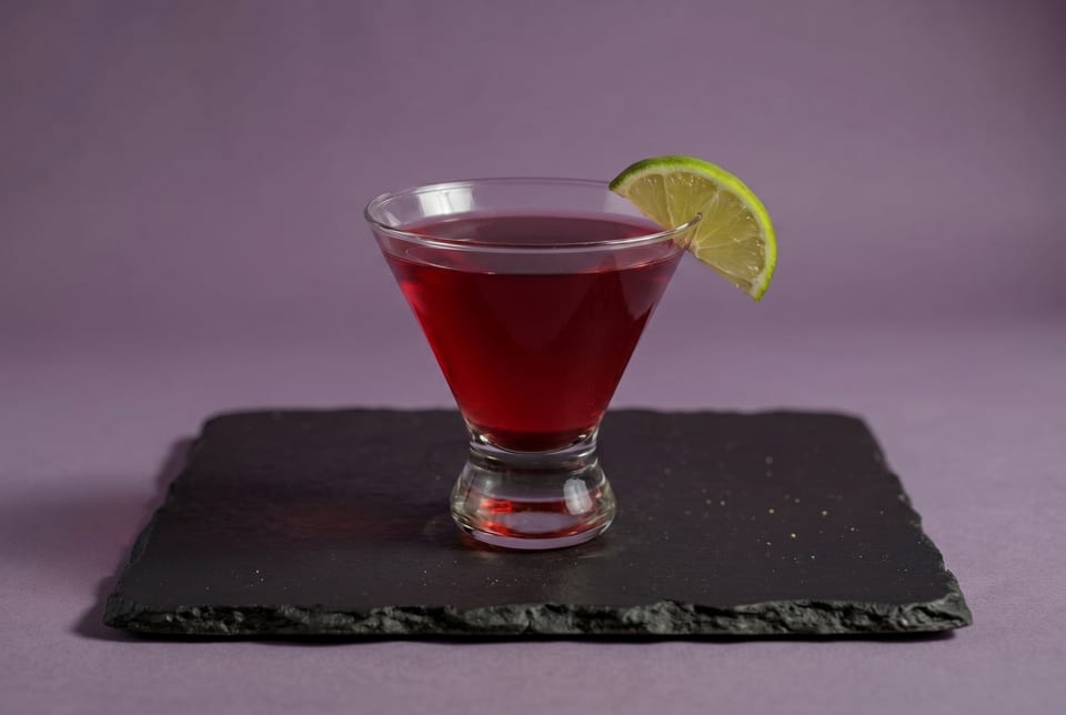 A red martini with a lime wedge garnish sits on a black slate coaster against a purple background.
