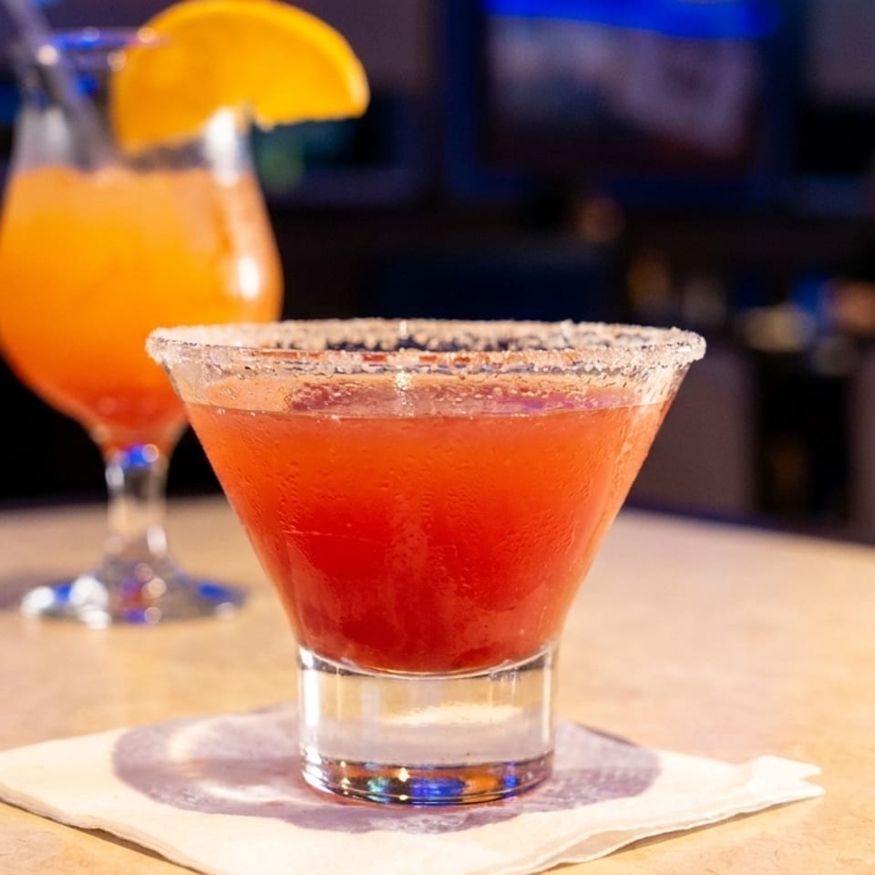 Two cocktails on a bar, one red with a salted rim, and one orange with an orange slice.