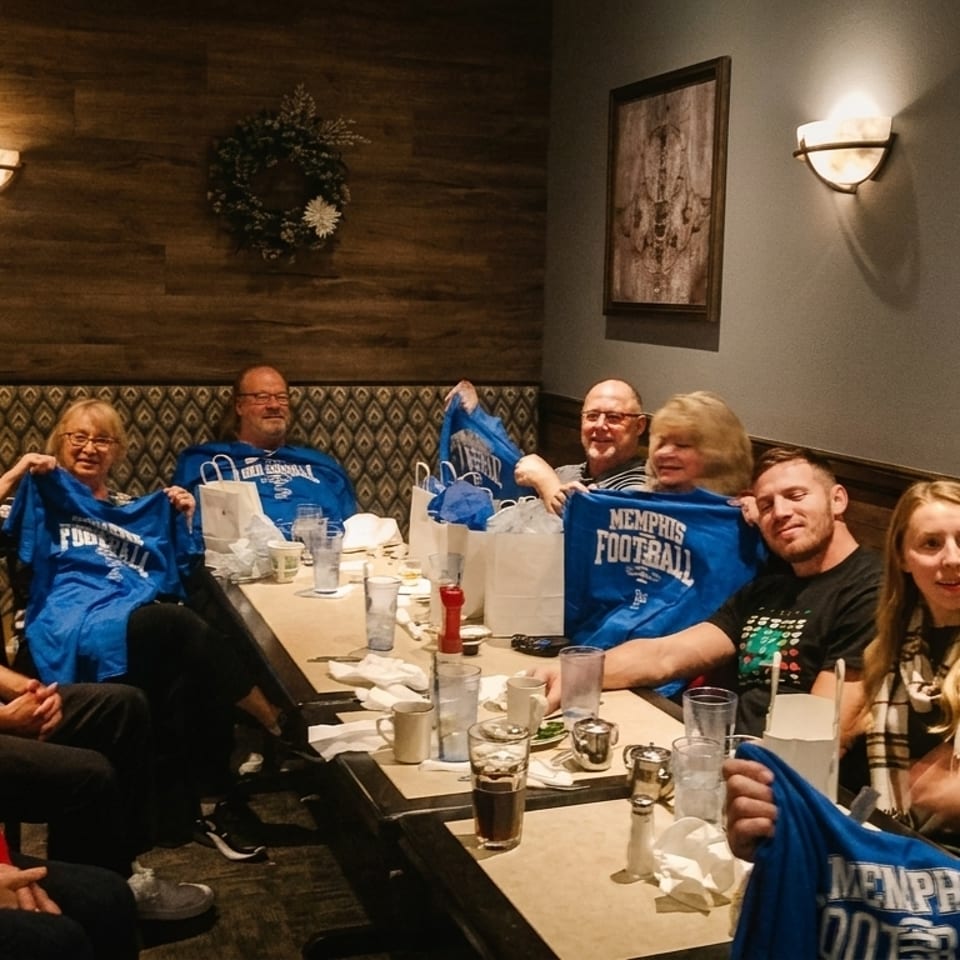 Group of people sitting at a long table in a restaurant, some holding up blue "Memphis Football" shirts.