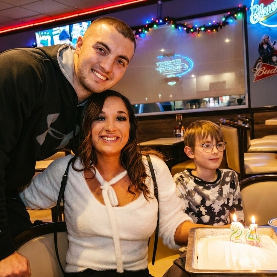 A family celebrates a birthday with cake and drinks at a restaurant with neon signs.